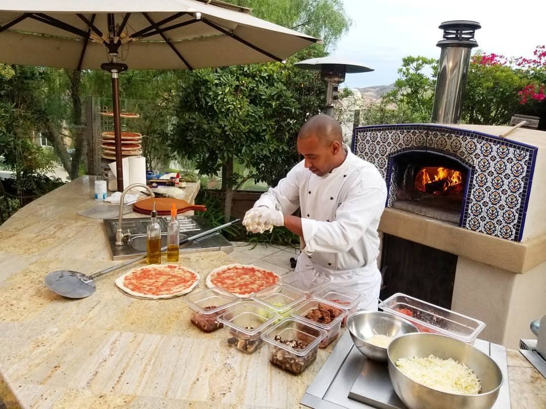 Chef topping fresh pizza dough beside a tiled outdoor oven at an Orange County catering event
